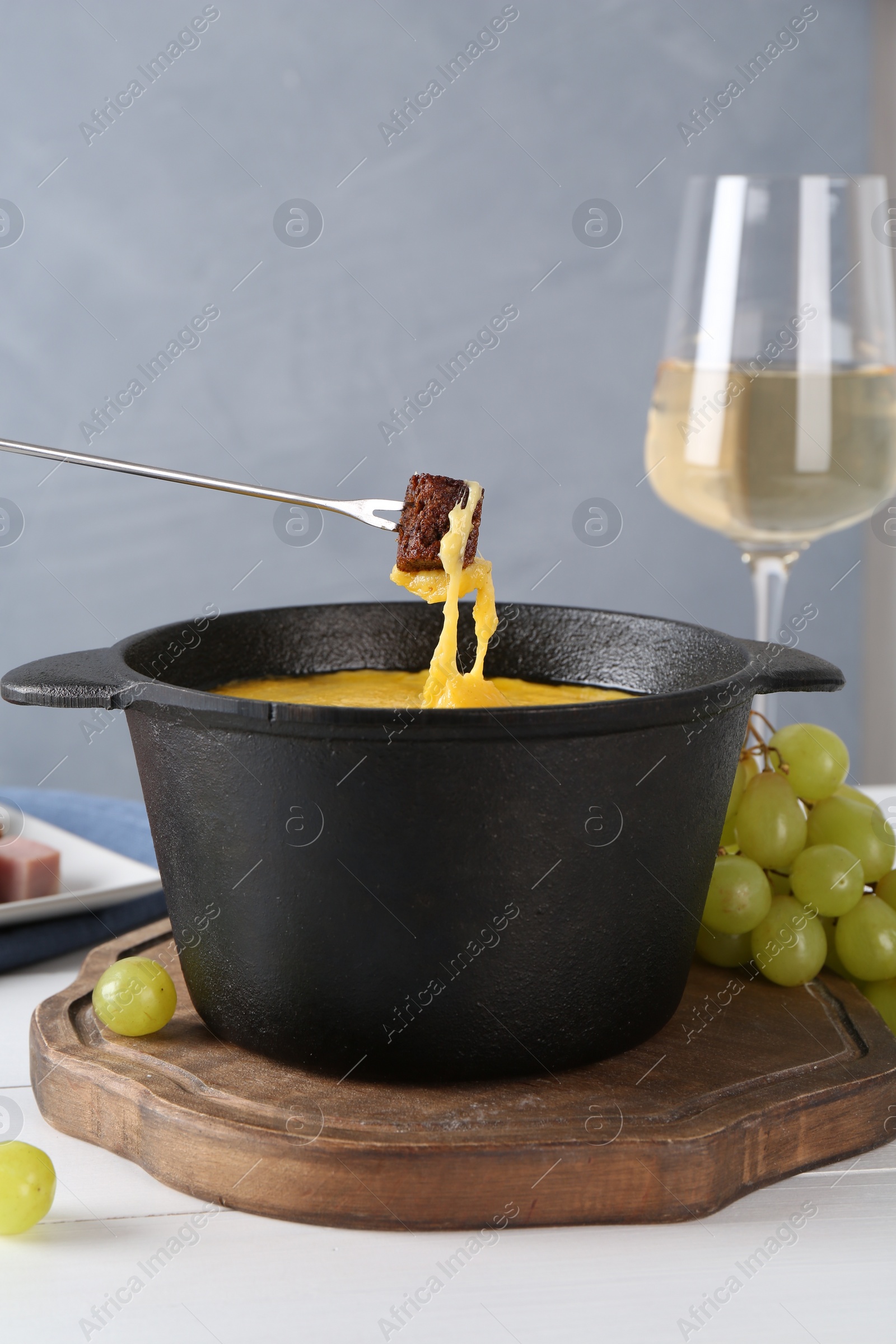 Photo of Dipping piece of bread into fondue pot with tasty melted cheese at white wooden table against gray background, closeup