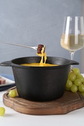 Photo of Dipping piece of bread into fondue pot with tasty melted cheese at white wooden table against gray background, closeup