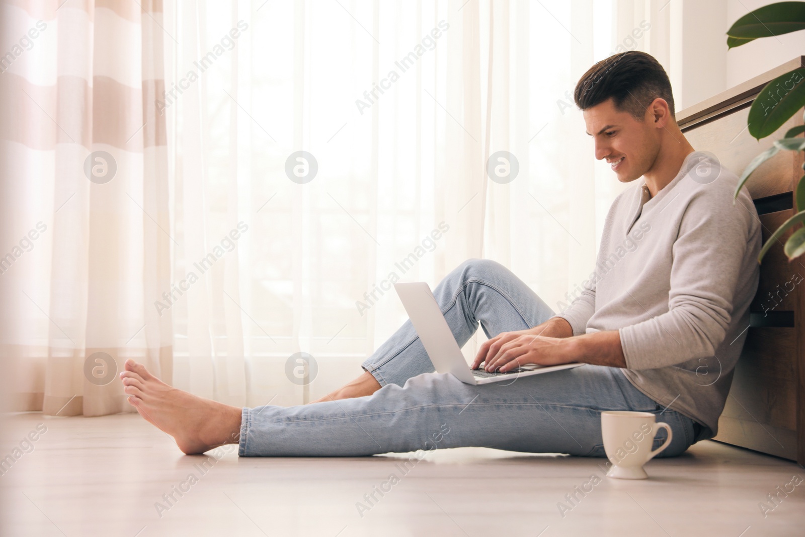 Photo of Man with cup of drink and laptop sitting on warm floor at home. Heating system