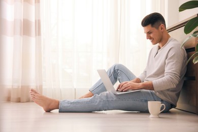 Man with cup of drink and laptop sitting on warm floor at home. Heating system