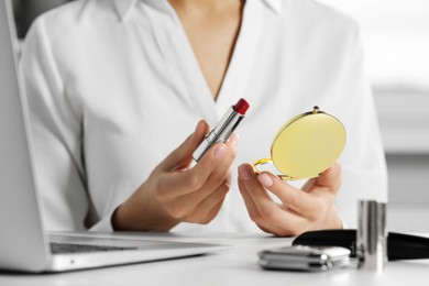 Photo of Young woman using cosmetic pocket mirror and lipstick at white table indoors, closeup