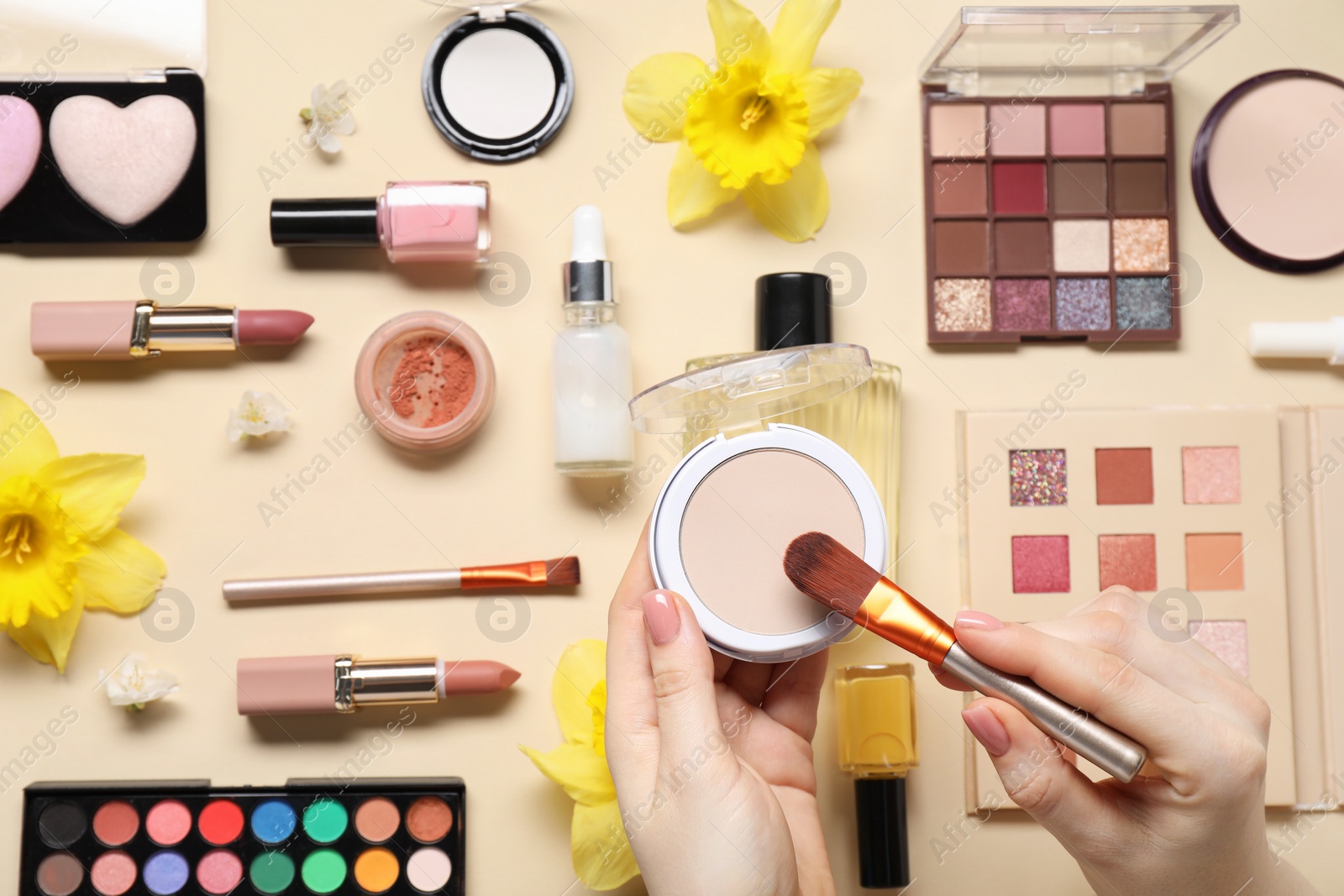 Photo of Woman applying face powder on brush, closeup. Different makeup products and beautiful spring flowers on beige background, top view