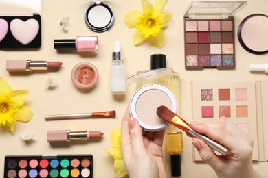 Photo of Woman applying face powder on brush, closeup. Different makeup products and beautiful spring flowers on beige background, top view