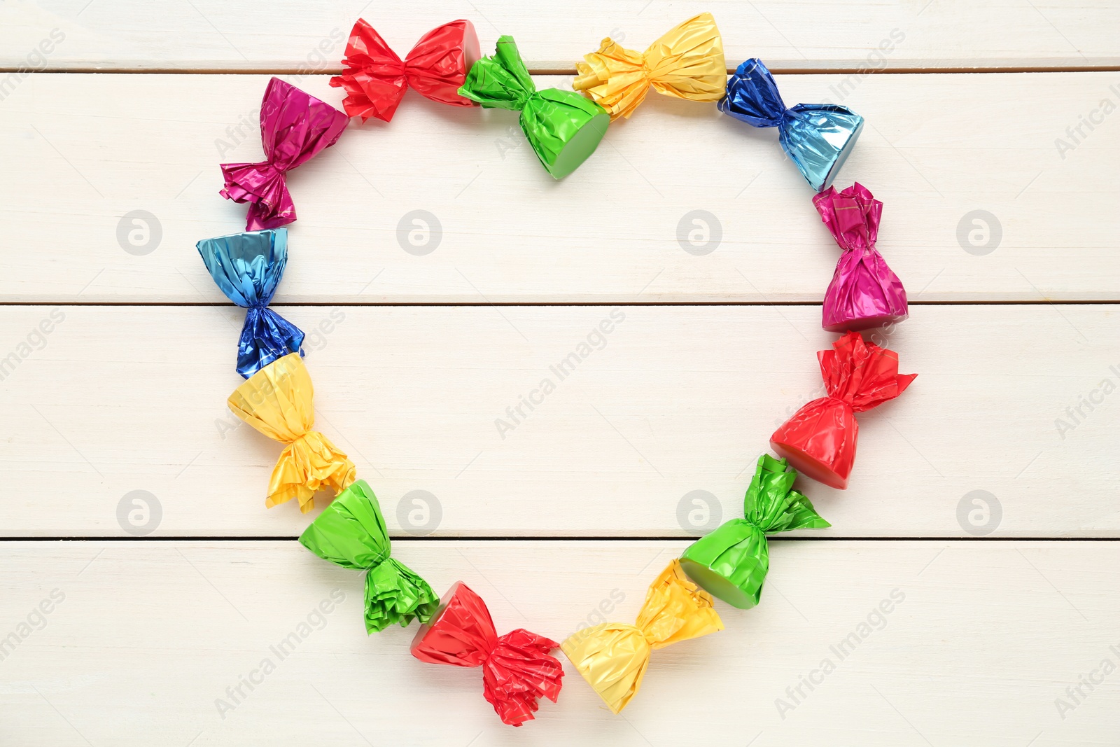 Photo of Heart shaped frame of candies in colorful wrappers on beige wooden table, flat lay. Space for text