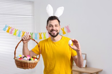Happy man in bunny ears headband holding wicker basket with painted Easter eggs indoors