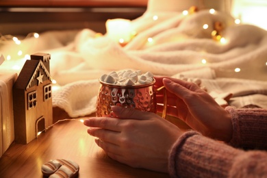 Photo of Woman holding cup of hot drink with marshmallows at wooden table, closeup. Magic Christmas atmosphere