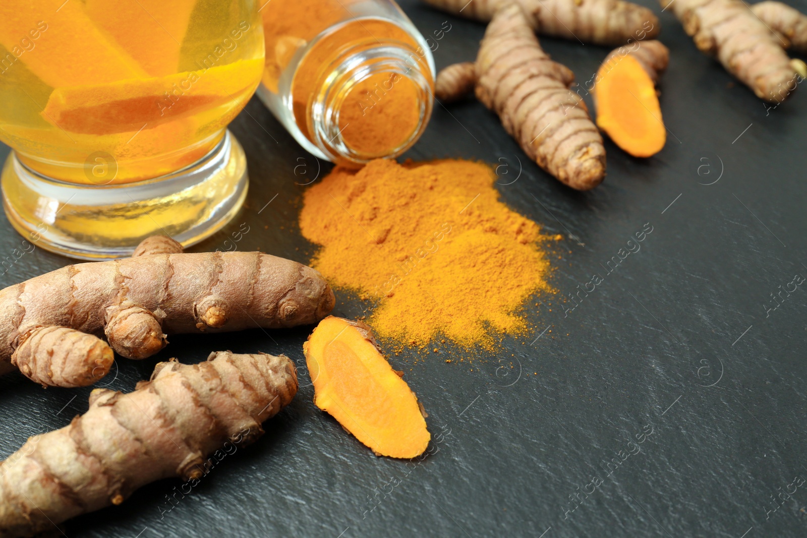 Photo of Glass cup of tasty tea, whole, cut turmeric roots and powder on black textured table, space for text