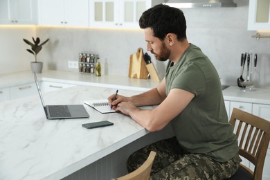 Photo of Soldier taking notes while working with laptop at white marble table in kitchen. Military service