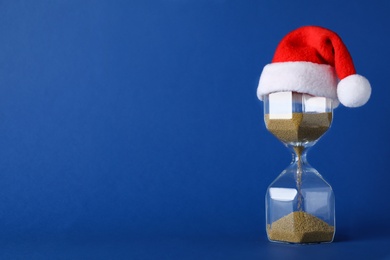 Photo of Hourglass and Santa hat on blue background, space for text. Christmas countdown