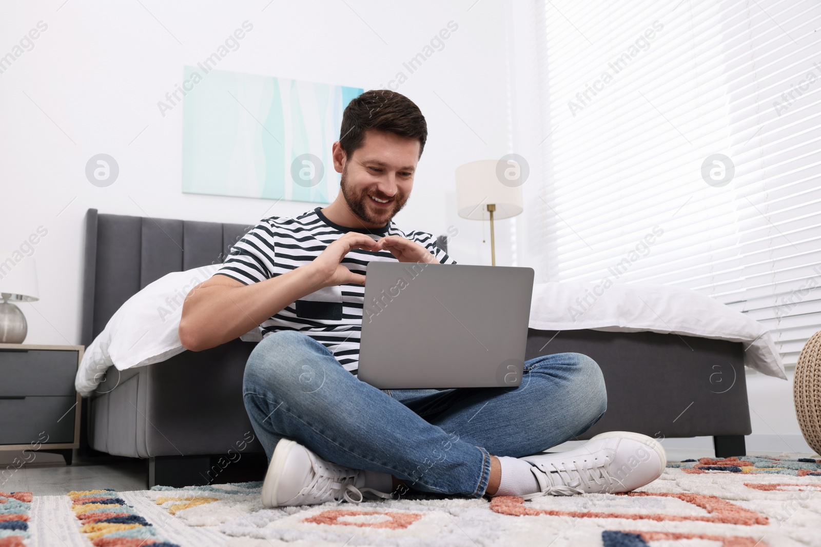 Photo of Happy man making heart with hands during video chat via laptop in bedroom. Long-distance relationship