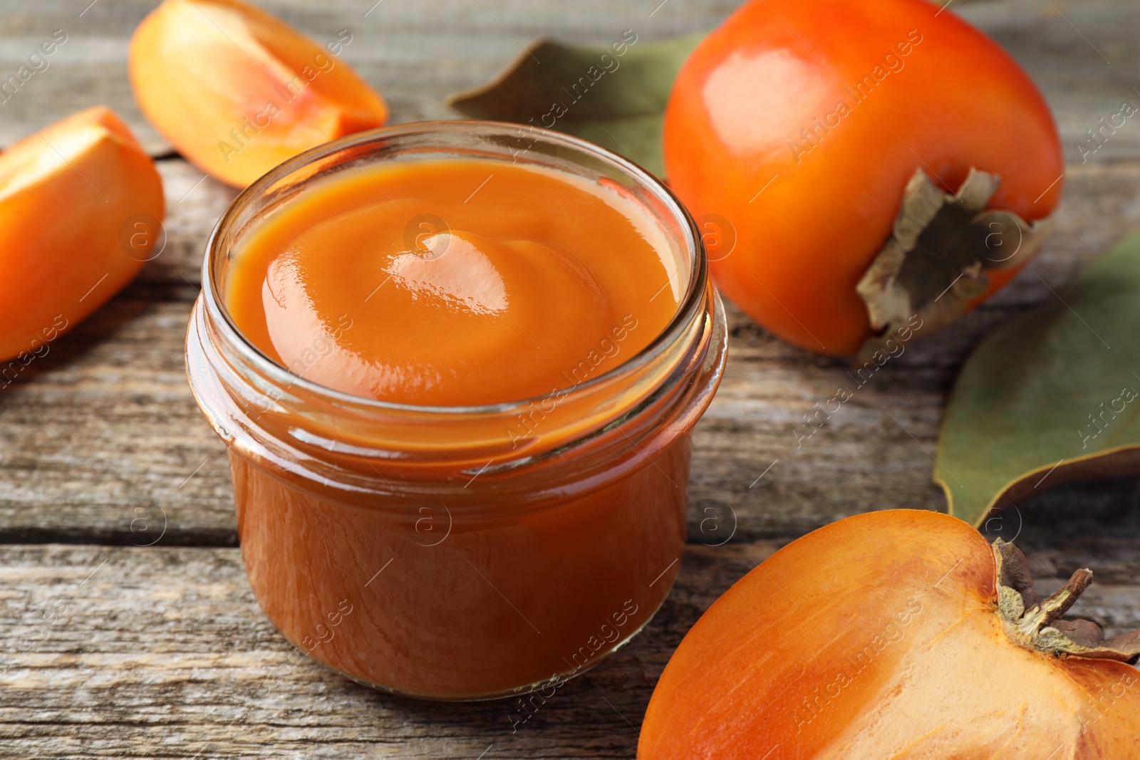 Photo of Delicious persimmon jam in glass jar and fresh fruits on wooden table, closeup