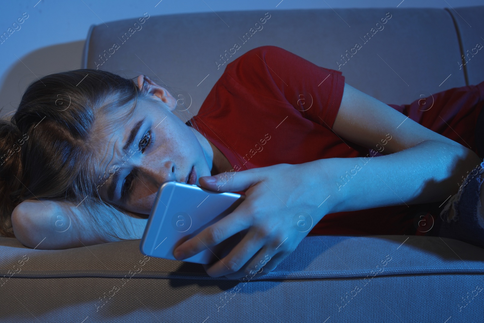 Photo of Upset teenage girl with smartphone lying on sofa in dark room. Danger of internet