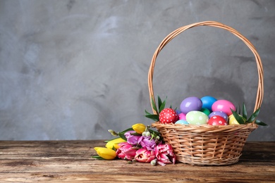 Photo of Wicker basket with bright painted Easter eggs and spring flowers on wooden table against grey background. Space for text