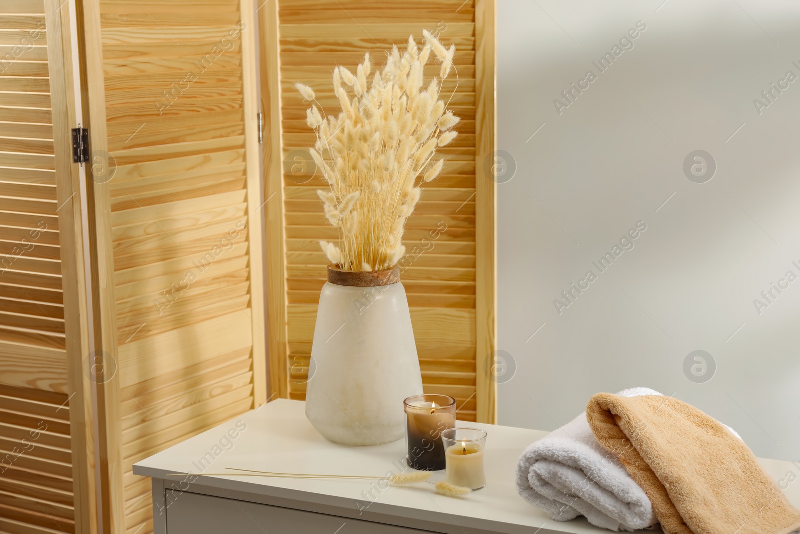 Photo of Vase with dried flowers, stacked towels and candles on chest of drawers in room. Interior design