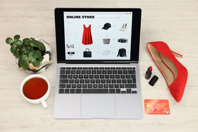 Photo of Online store. Laptop with open website, credit card, red high-heeled shoe and lipstick on wooden surface, above view