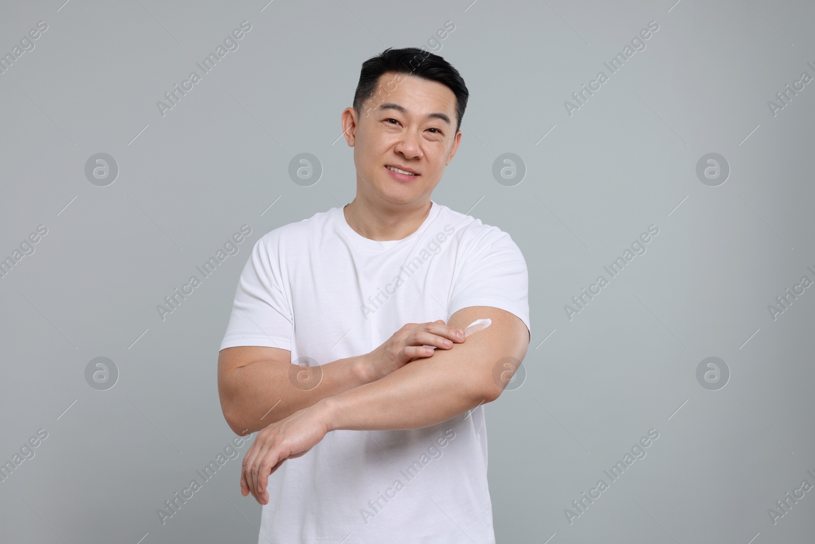 Photo of Handsome man applying body cream onto his arm on light grey background