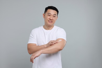 Handsome man applying body cream onto his arm on light grey background