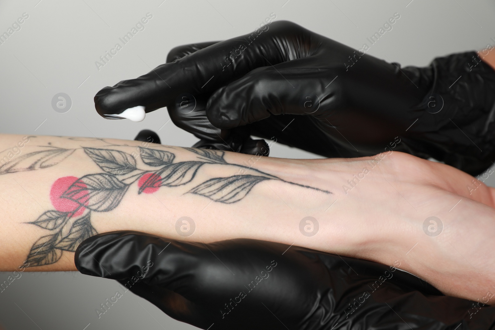 Photo of Worker in gloves applying cream on woman's arm with tattoo against light background, closeup