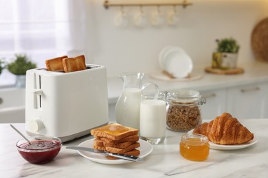 Photo of Making toasts for breakfast. Appliance, crunchy bread, honey, jam, milk and croissant on white marble table in kitchen