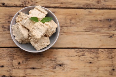 Photo of Bowl with pieces of tasty halva and mint leaves on wooden table, top view. Space for text