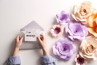 Apology. Woman holding envelope and card with phrase I Am Sorry near beautiful flowers at white table, top view