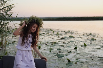 Photo of Cute little girl wearing wreath made of beautiful flowers in wooden boat at sunset
