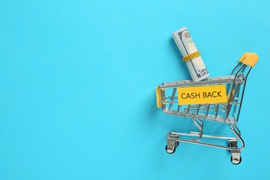 Card with word Cashback, rolled dollar banknotes and shopping cart on light blue background, flat lay. Space for text