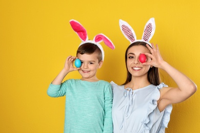 Mother and son in bunny ears headbands holding Easter eggs near eyes on color background