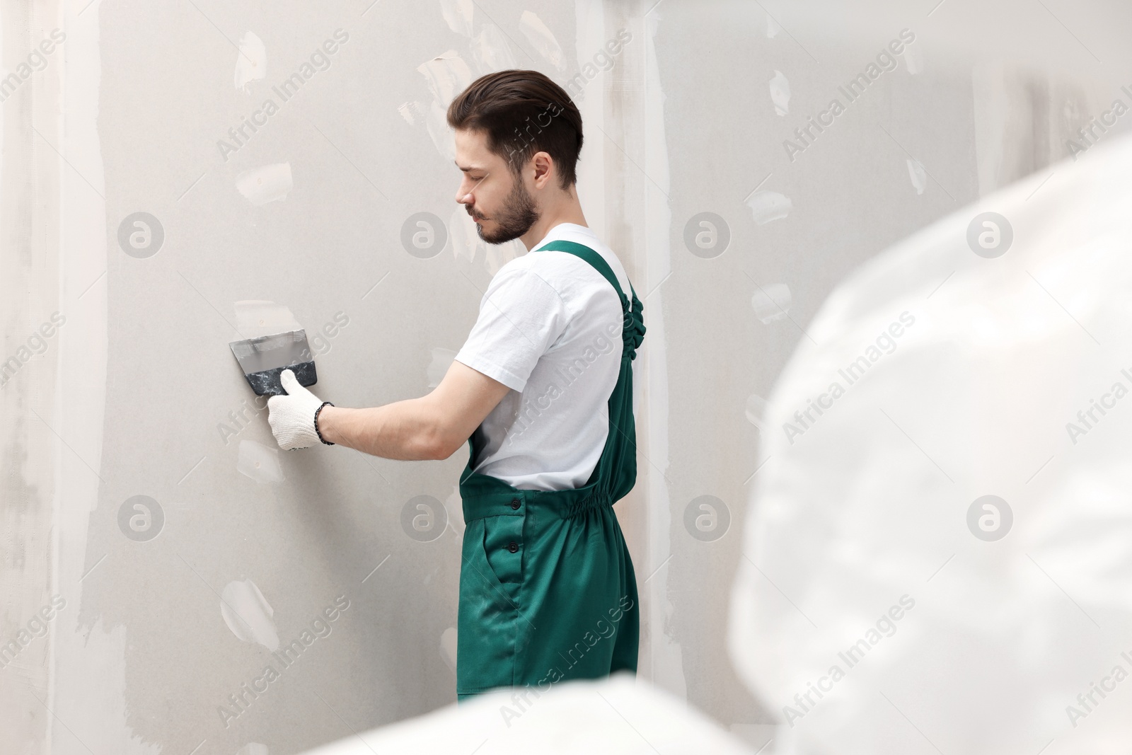 Photo of Worker in uniform plastering wall with putty knife indoors