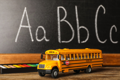 Photo of School bus model and stationery on wooden table near chalkboard. Transport for students