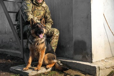 Photo of Ukrainian soldier with German shepherd dog sitting outdoors, closeup. Space for text