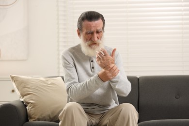 Photo of Senior man suffering from pain in hand on sofa at home. Rheumatism symptom