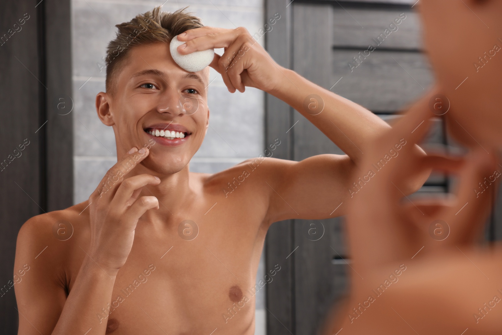 Photo of Happy young man washing his face with sponge near mirror in bathroom