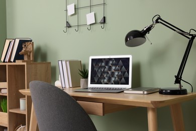 Photo of Modern laptop, books, lamp and stationery on wooden desk near green wall. Home office