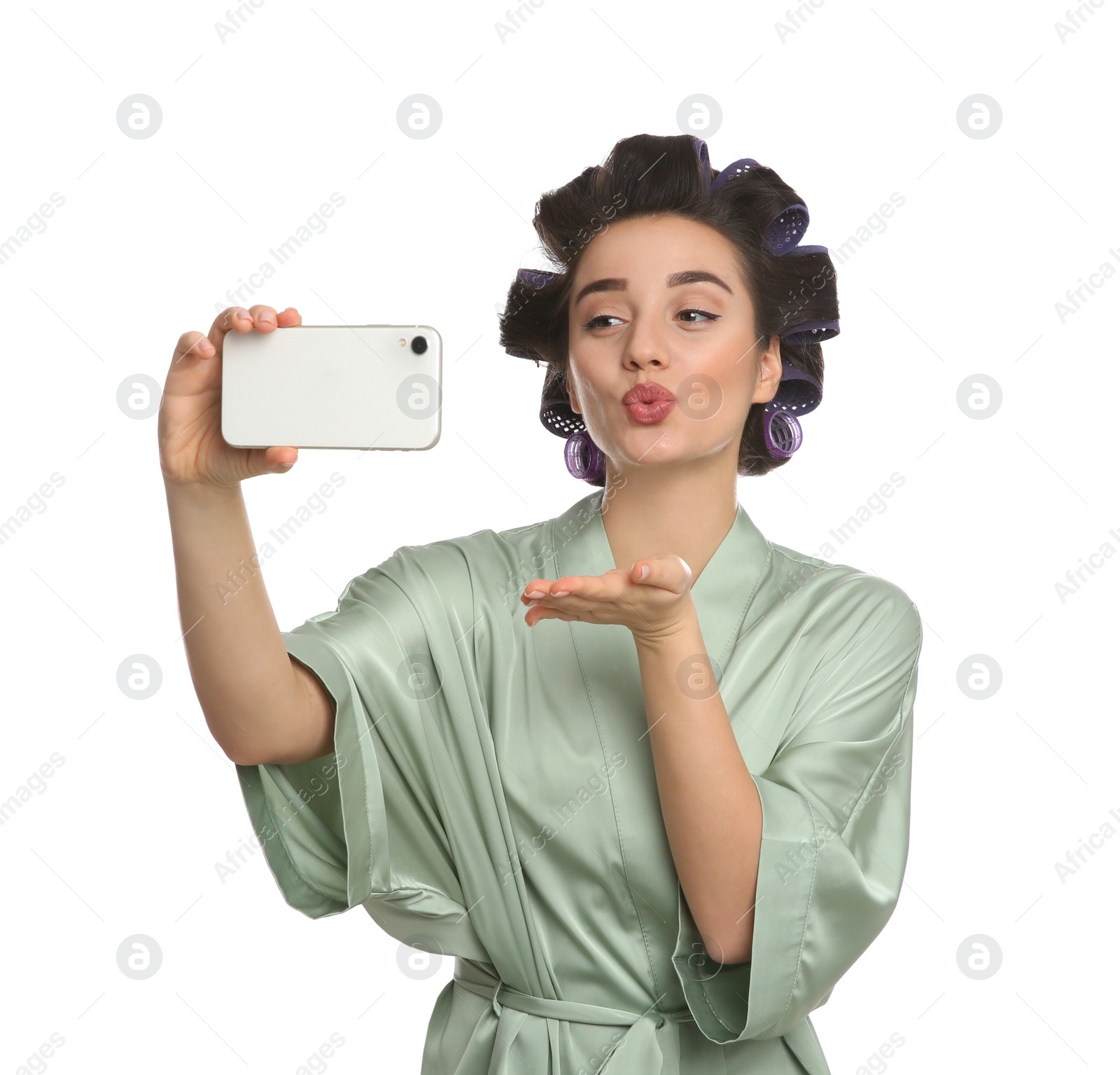 Photo of Young woman in silk bathrobe with hair curlers taking selfie on white background