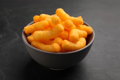 Many tasty cheesy corn puffs in bowl on black table, closeup view