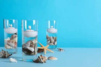 Photo of Burning candles in glass holders with seashells on light blue background, space for text