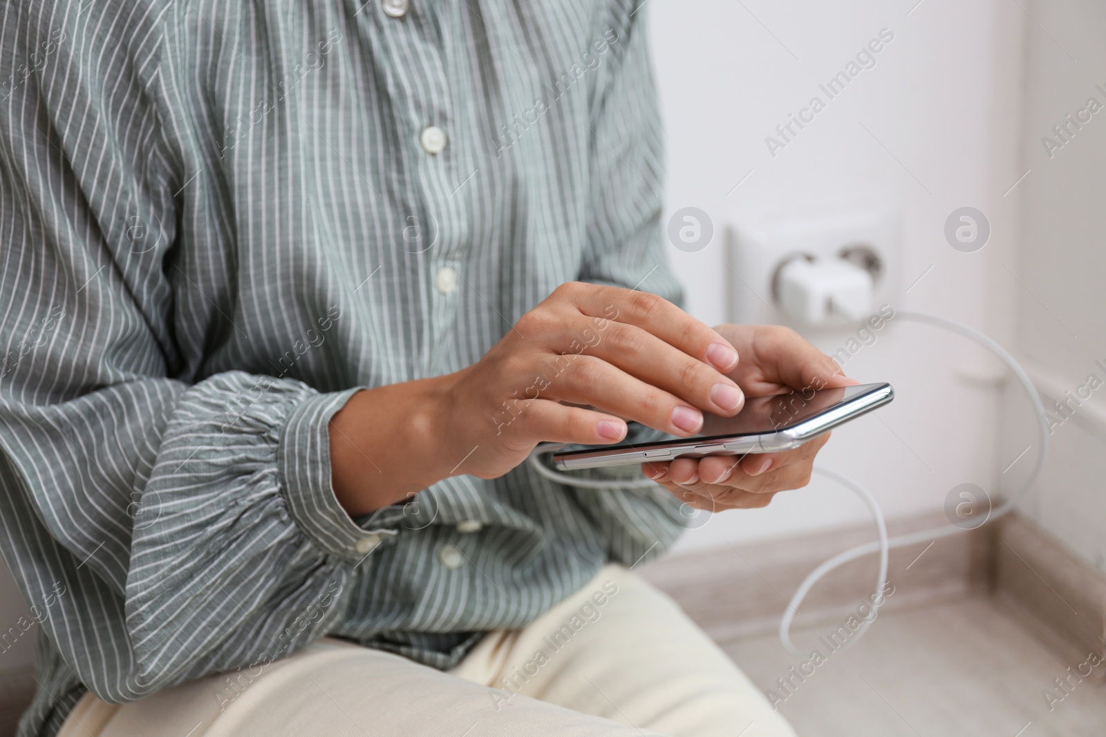 Photo of Woman using mobile phone with connected charger near white brick wall indoors, closeup