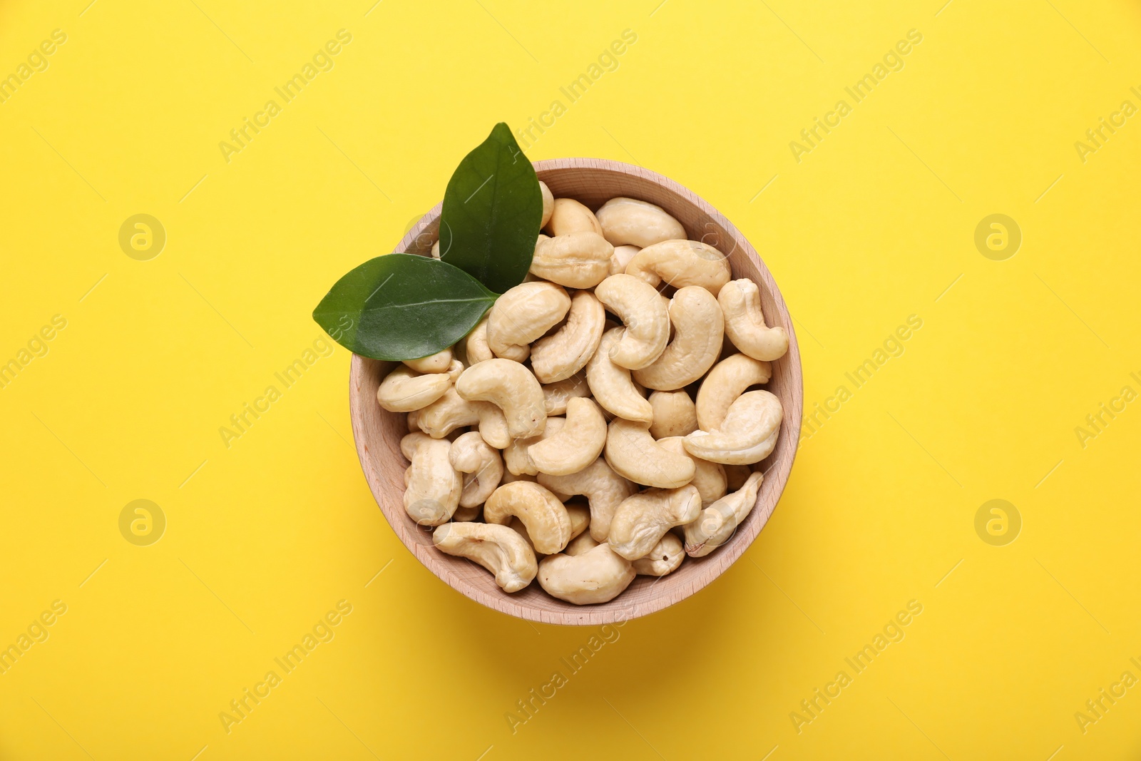 Photo of Tasty cashew nuts and green leaves in bowl on yellow background, top view