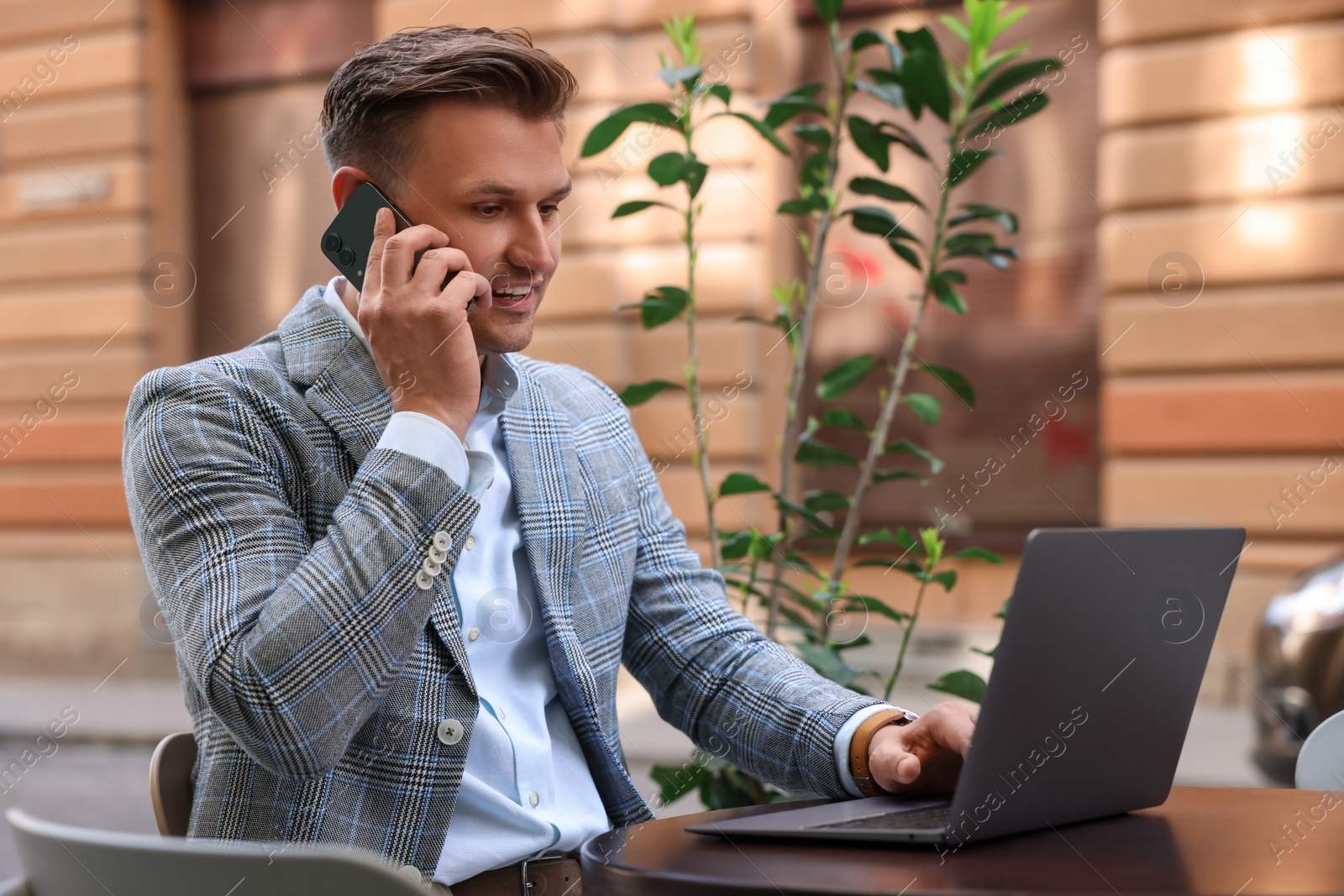 Photo of Handsome man talking on smartphone while using laptop at table in outdoor cafe