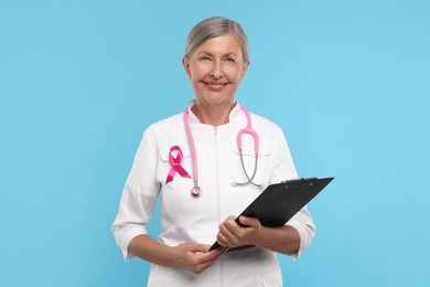 Photo of Doctor with pink ribbon, clipboard and stethoscope on light blue background. Breast cancer awareness