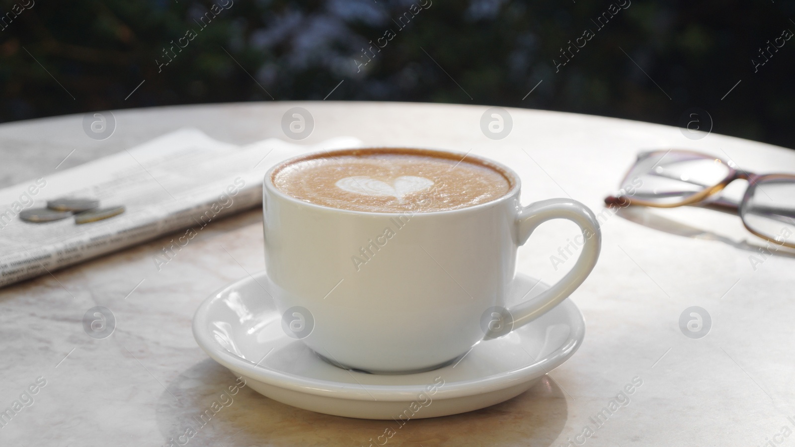 Photo of Cup of delicious coffee, glasses and newspaper on beige marble table outdoors
