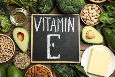 Photo of Small chalkboard with phrase Vitamin E and different products on wooden table, flat lay