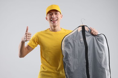 Photo of Dry-cleaning delivery. Happy courier holding garment cover with clothes and showing thumbs up on light grey background