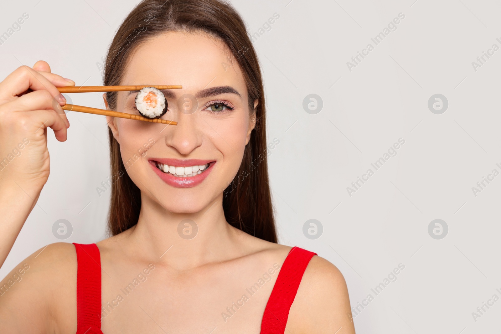 Photo of Funny young woman holding sushi roll with chopsticks on light background. Space for text