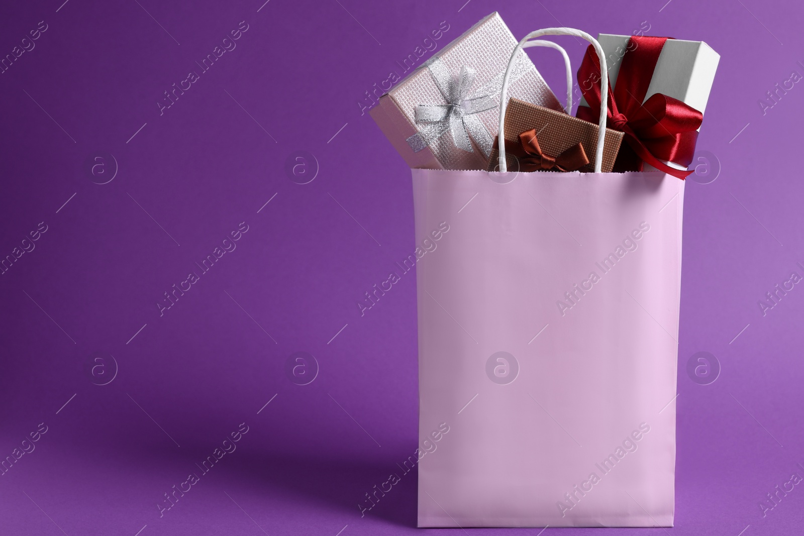 Photo of Pink paper shopping bag full of gift boxes on purple background. Space for text