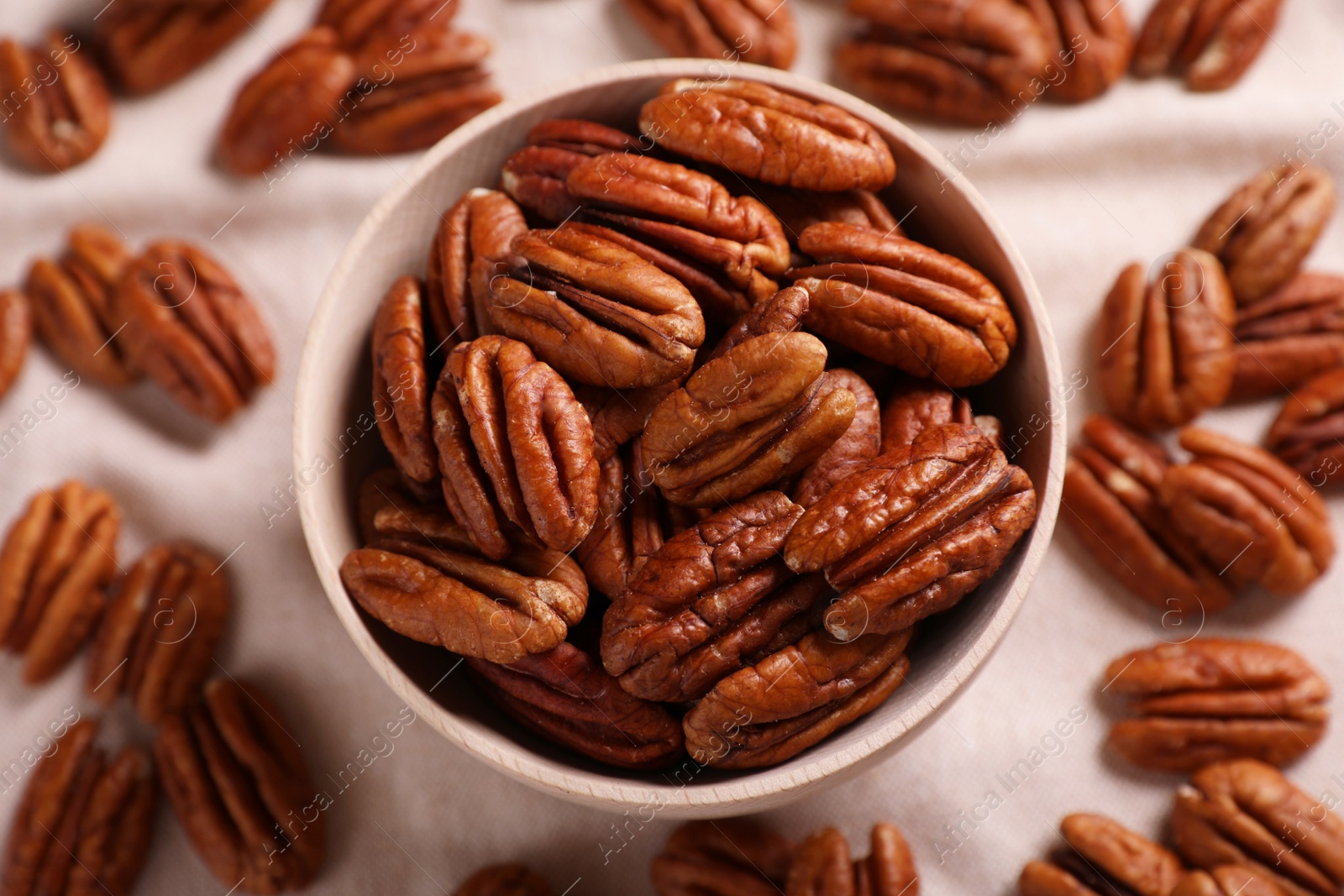 Photo of Tasty pecan nuts with bowl on beige cloth, flat lay