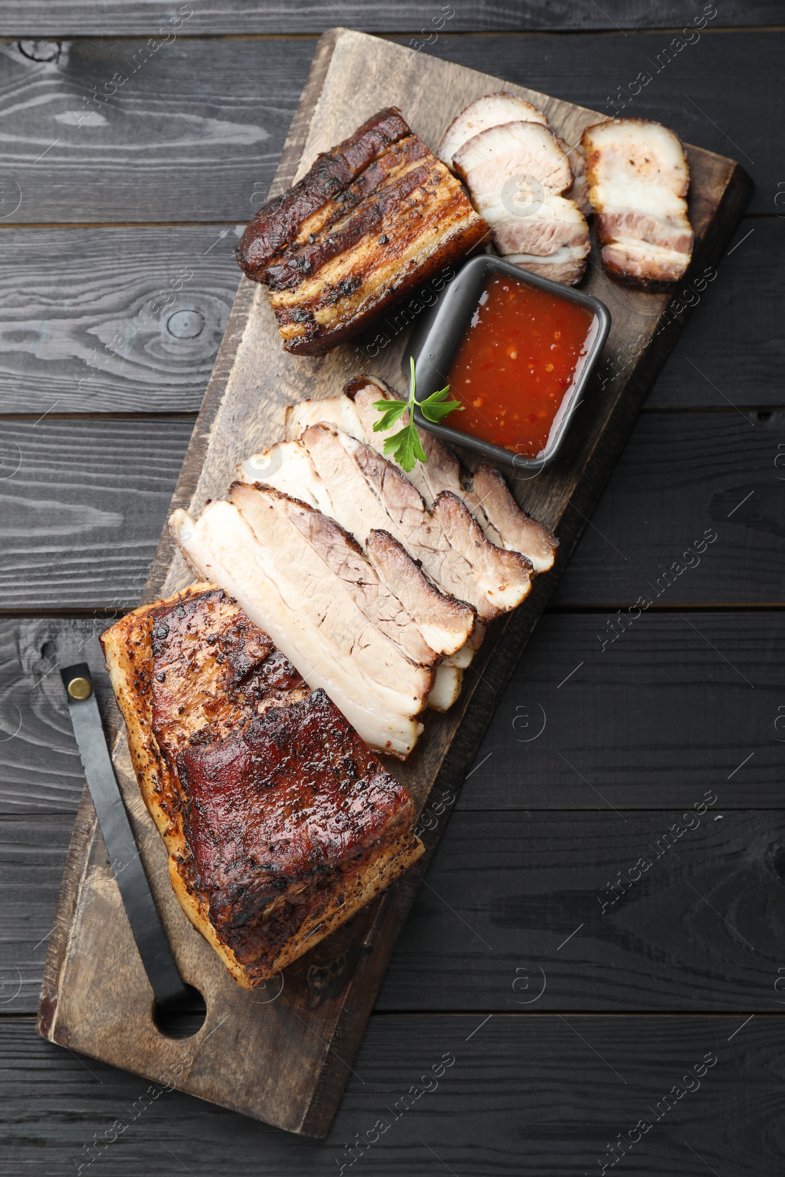 Photo of Pieces of baked pork belly served with sauce and parsley on black wooden table, top view