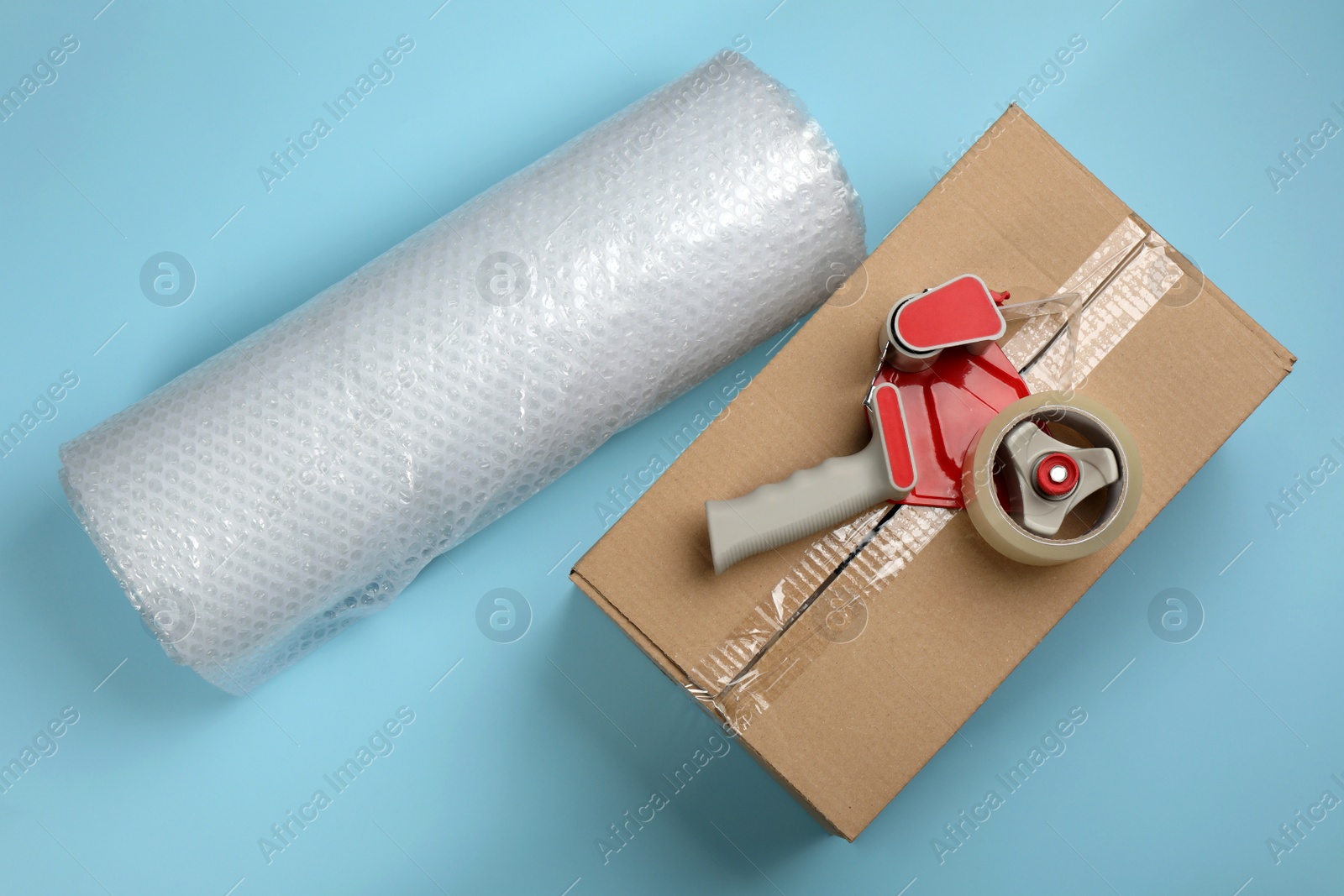 Photo of Bubble wrap roll, cardboard box and tape dispenser on turquoise background, flat lay