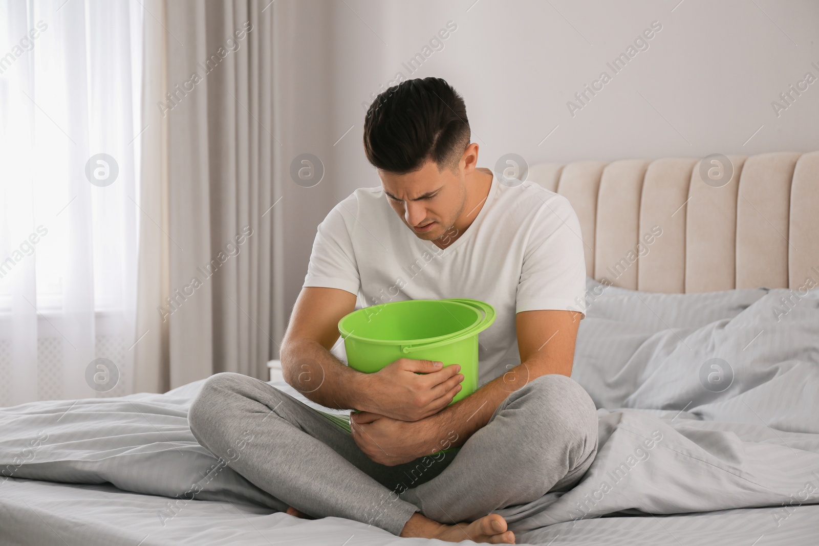 Photo of Man with bucket suffering from nausea on bed at home. Food poisoning